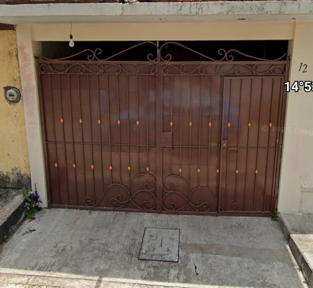 A brown wrought iron gate with decorative elements is displayed, providing security and access to the property. The gate is set within a concrete wall, with a clear view of the driveway area. A street number is visible on the side.