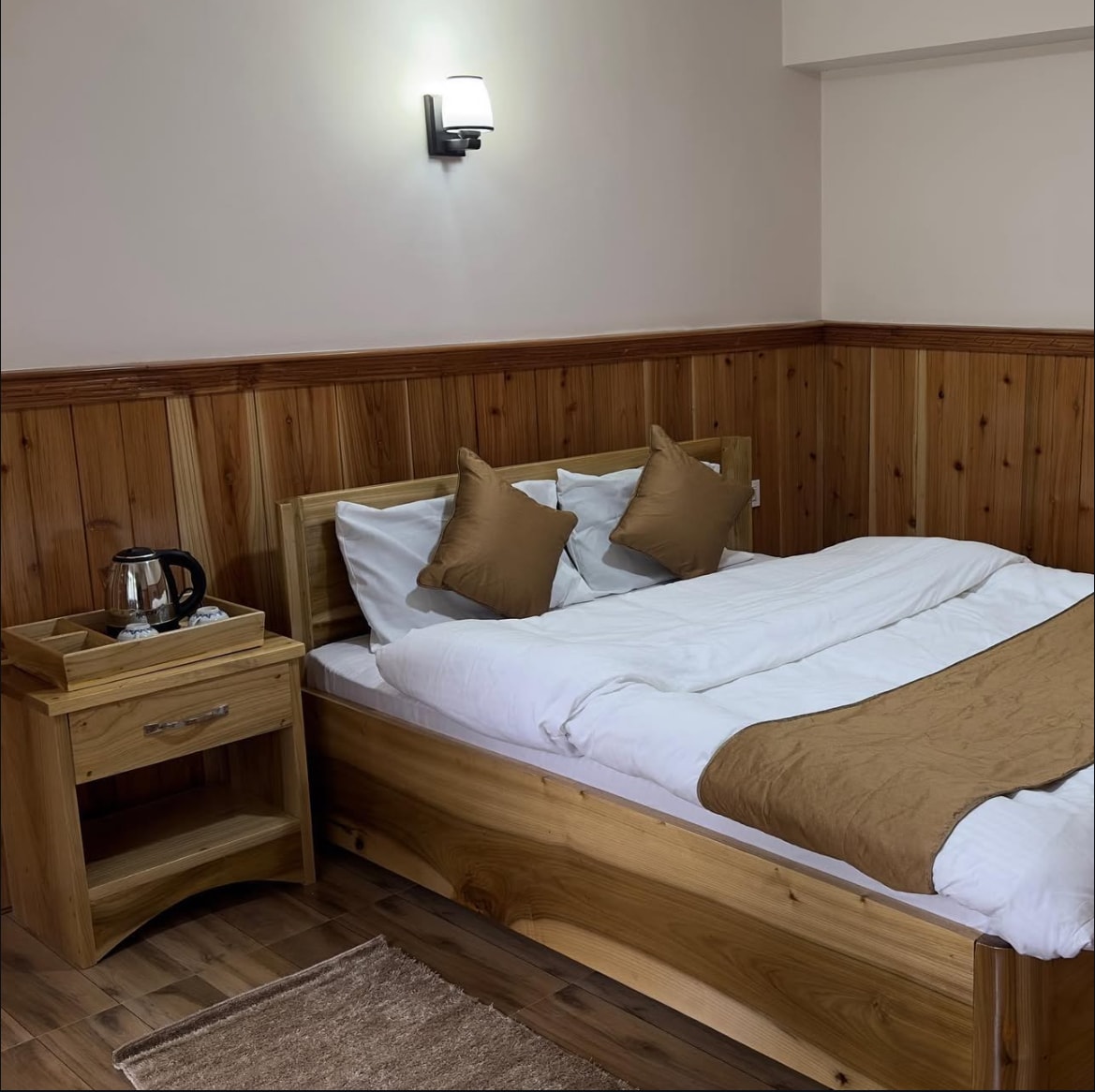 A cozy bedroom features wooden paneling and a spacious bed adorned with beige and white bedding. Two decorative pillows complement the bedding. A brown nightstand holds a kettle, and soft lighting is provided by a wall-mounted sconce.