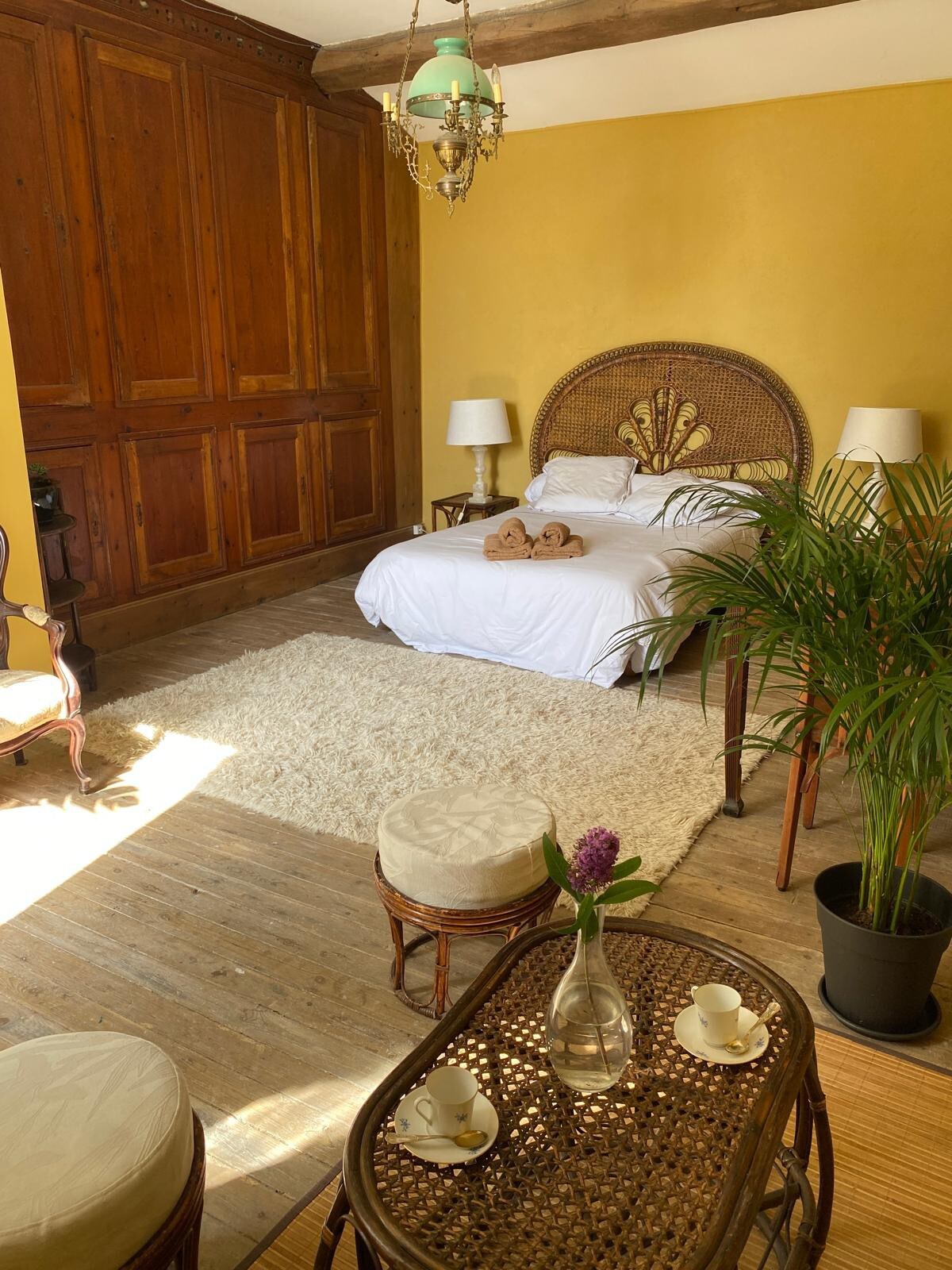 A cozy bedroom features a spacious bed set against a warm yellow wall. Natural light floods the room, highlighting a textured rug and wicker furniture. A small table with two cups sits next to a decorative plant, adding a touch of greenery to the inviting space.