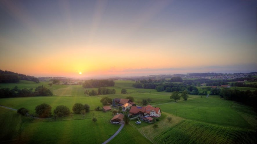 A scenic view captures the expansive landscape at sunset, with the sun rising over rolling green fields. A collection of buildings is visible in the foreground, surrounded by trees and open spaces, creating a serene countryside atmosphere.