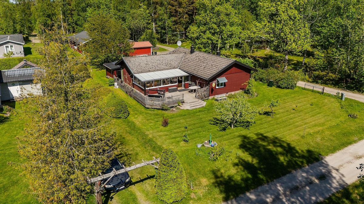 An aerial view shows a spacious red cabin surrounded by lush greenery, with a large deck featuring a covered area. The well-maintained lawn is dotted with trees and garden features. A peaceful dirt path leads toward the cabin, emphasizing its tranquil setting.