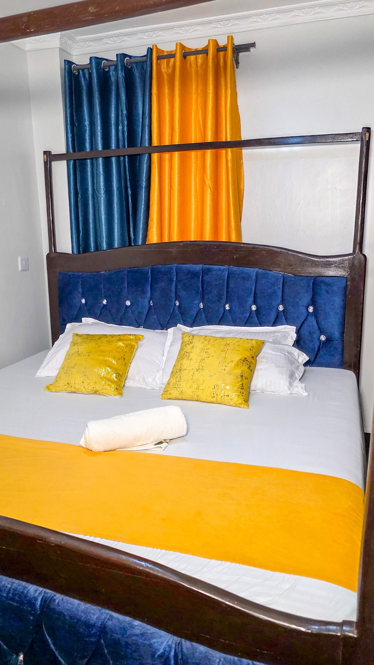 A cozy bedroom features a plush bed with a tufted blue headboard and crisp white linens. Yellow and blue curtains frame a window, while yellow decorative pillows add a touch of color. A rolled towel is placed on a bright yellow blanket, enhancing the welcoming ambiance.