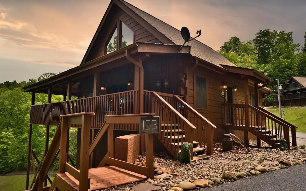 The cabin features a rustic wooden exterior with a sloped roof and several steps leading up to the entrance. An inviting front porch is enhanced by railings, and natural landscaping includes stones and greenery surrounding the foundation.
