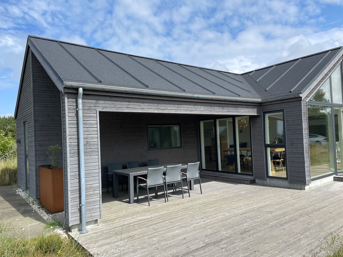 A modern summer house is presented with a dark timber exterior and a slanted roof. An outdoor dining area featuring a large table and several chairs is situated on a wooden deck. Large glass doors provide views into the interior living space.