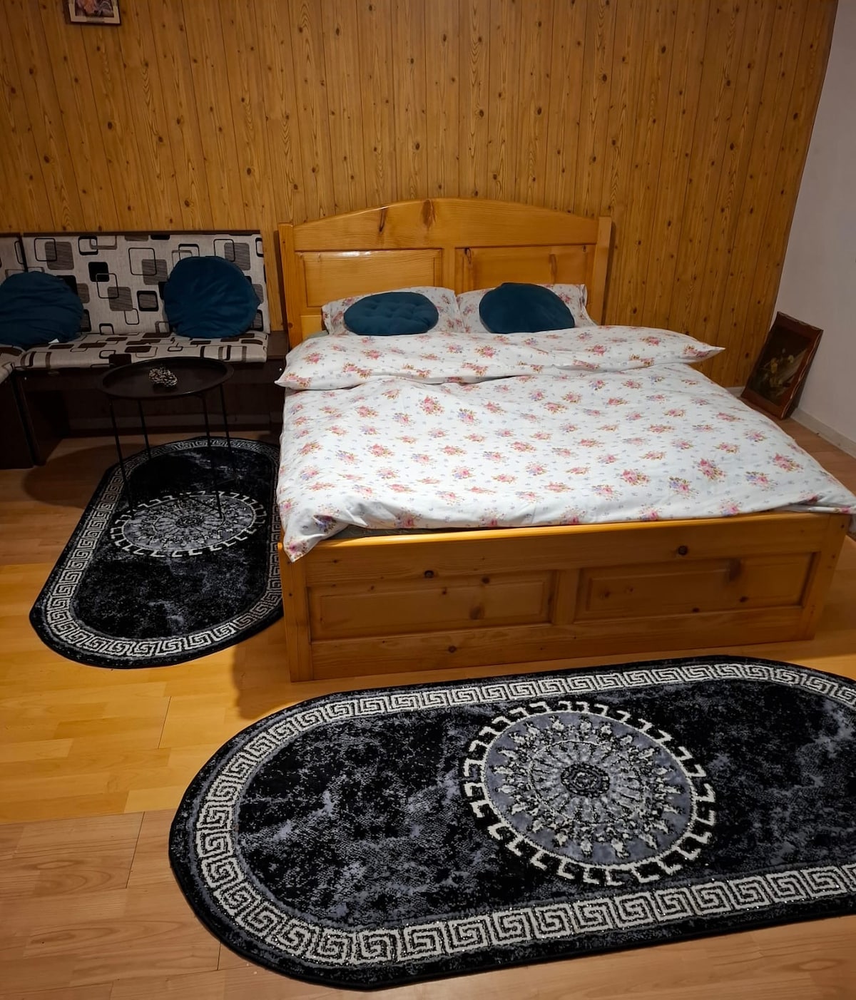 A sleeping area features a wooden bed with floral bedding and two decorative pillows. A cozy seating arrangement is visible in the corner, accompanied by a small black table. Dark rugs with intricate designs are placed on the wooden floor, enhancing the room's comfort.