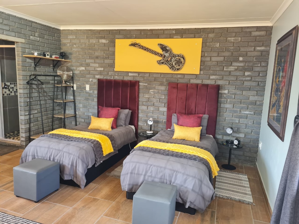The bedroom features two individual beds with dark headboards, arranged against a textured gray brick wall. Vibrant yellow and purple pillows add color to the neutral bedding. A piece of wall art depicting a guitar hangs above, while bedside lamps provide illumination on either side.