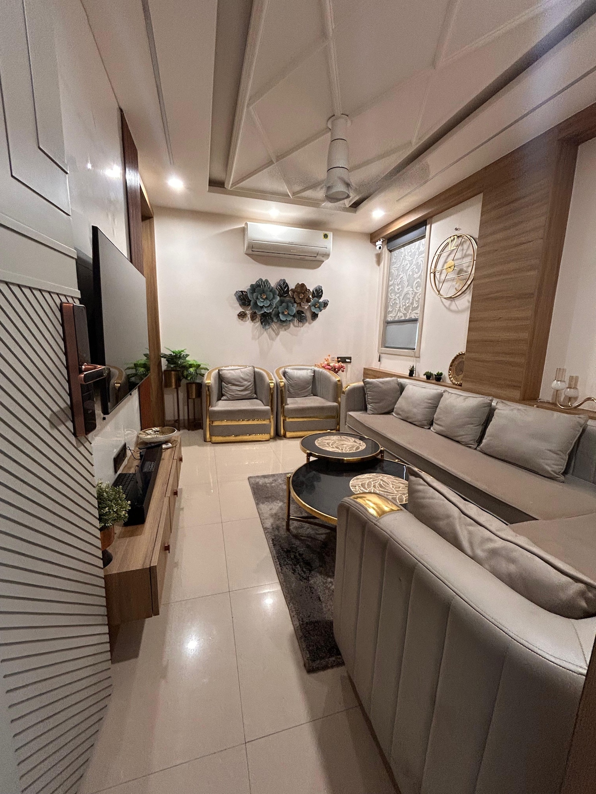A stylish living area features a comfortable sectional sofa and four armchairs, surrounded by soft lighting. A round coffee table rests at the center, adding functionality. Wall decor includes an artistic display, while plants enhance the overall ambiance.