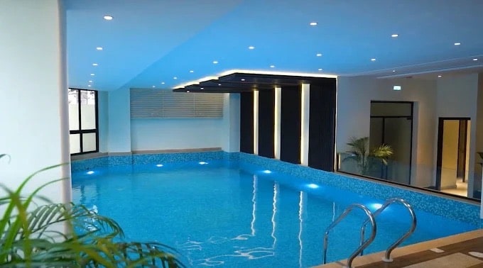 An indoor infinity pool is featured, surrounded by sleek walls and modern lighting. Clear blue water reflects ambient lights, creating a serene atmosphere. Lush greenery is positioned at the edges, adding an element of nature to the space. Steps lead into the pool from a stone deck.