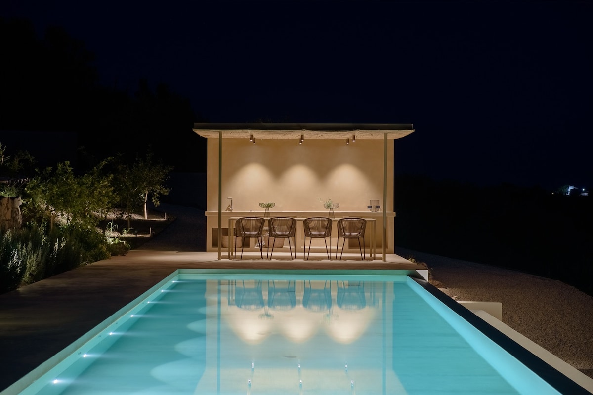 A tranquil evening scene features a well-lit outdoor bar area adjacent to a glimmering pool. The reflection of soft lighting on the water enhances the pool's inviting atmosphere, while the surrounding landscape is subtly illuminated, creating a serene ambiance.