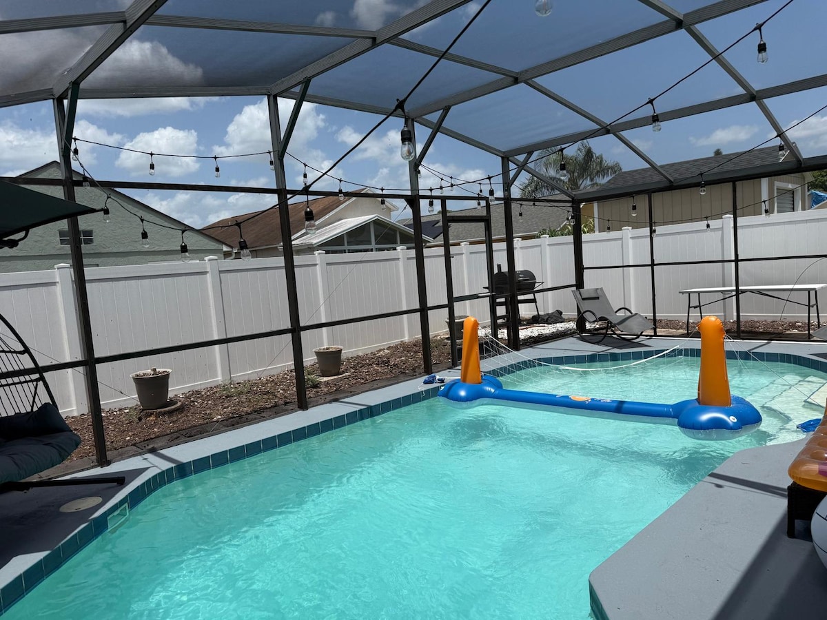 A private swimming pool is enclosed by a screened structure, featuring a light blue pool float and surrounded by potted plants. Comfortable lounge chairs are positioned alongside the pool under a shaded area, with decorative string lights overhead.