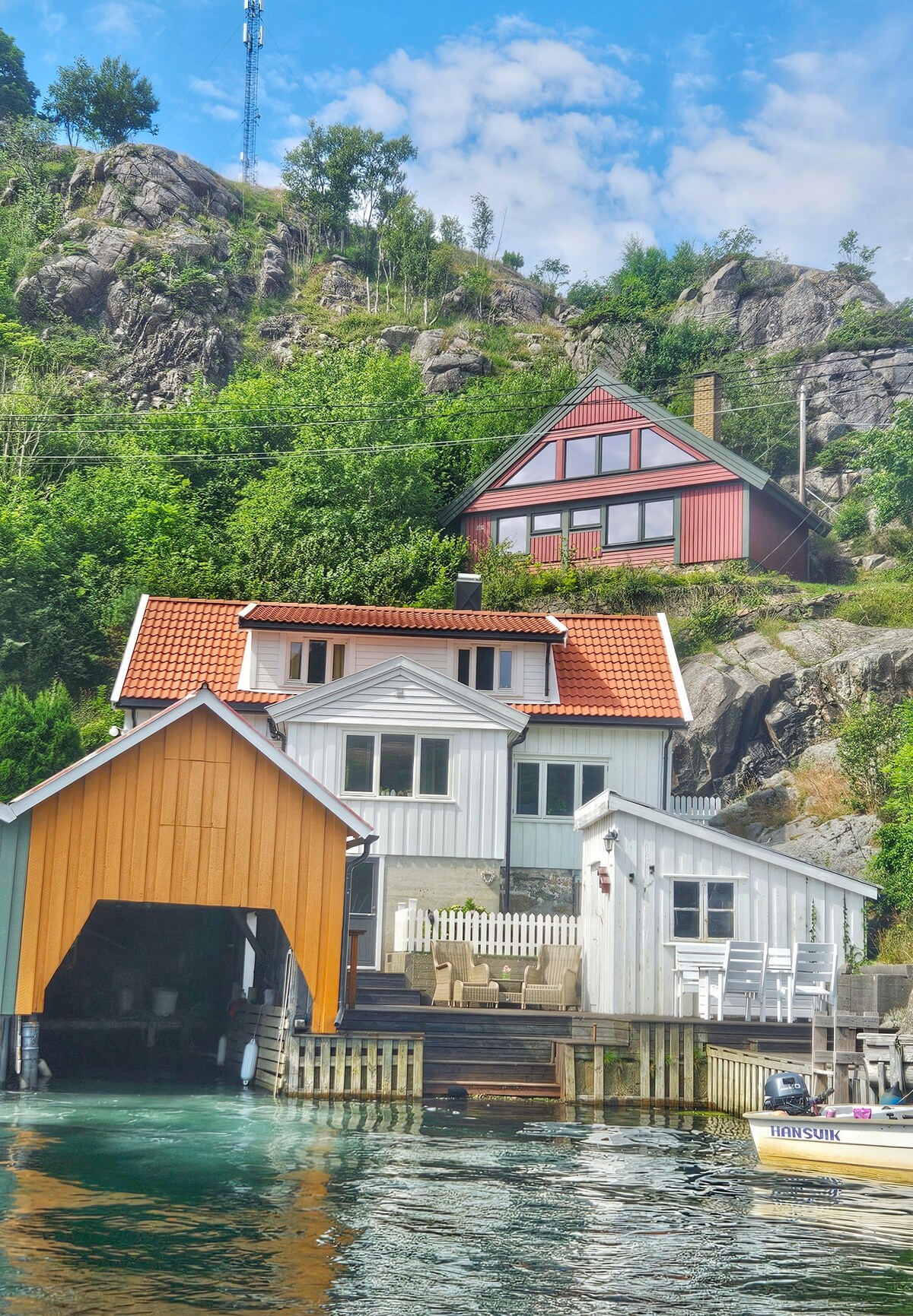 The charming three-story house is positioned by the seaside, featuring distinct colored facades with a red roof and orange garage. A spacious terrace with seating is visible, alongside the water, accentuated by the lush greenery of surrounding hills and a rocky backdrop.
