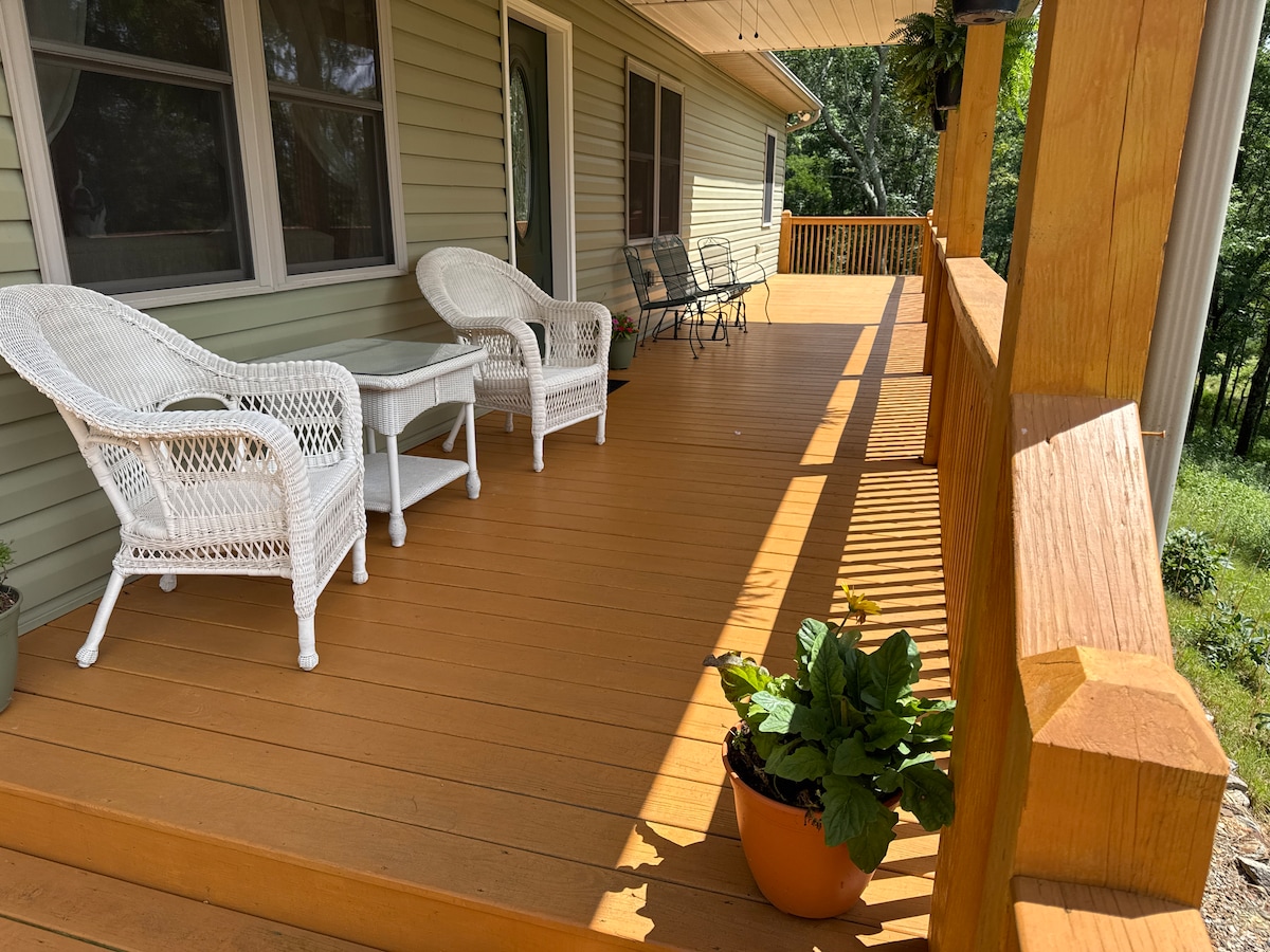 The spacious front porch offers a relaxing outdoor space with two white wicker chairs and a small table, arranged on wooden flooring. A decorative plant sits in a pot, adding a touch of greenery, while sunlight casts soft shadows across the area.