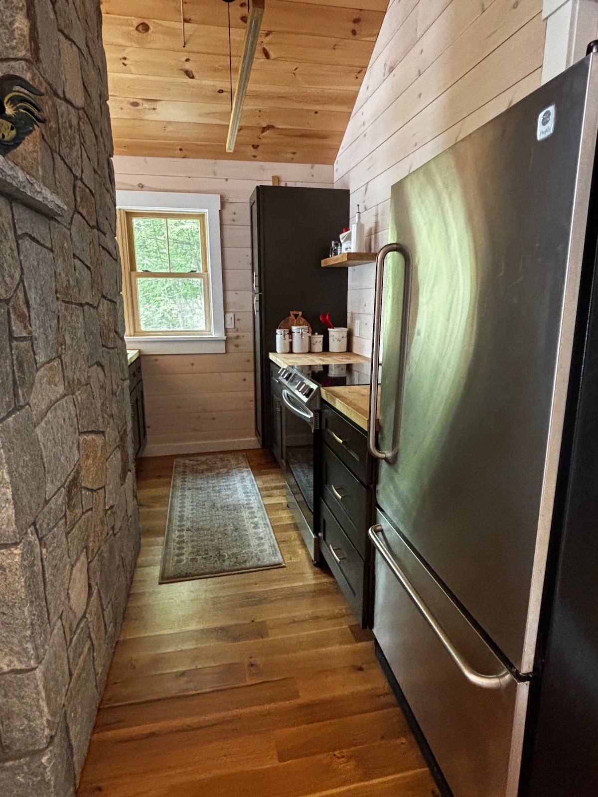 A narrow kitchen space is highlighted by wooden walls and stone accents. Stainless steel appliances, including a refrigerator and stove, are positioned against the dark cabinetry. A textured area rug extends along the floor, and natural light enters through a window, creating a bright atmosphere.