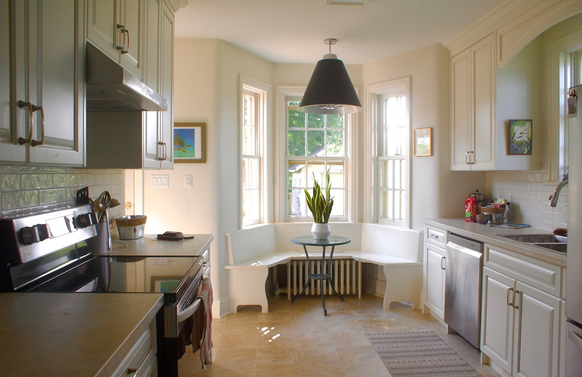 A bright kitchen is showcased with white cabinetry and stainless steel appliances. A cozy bay window nook features a built-in bench surrounding a small table, while natural light illuminates the space. Ceramic tiles add a warm touch to the flooring.