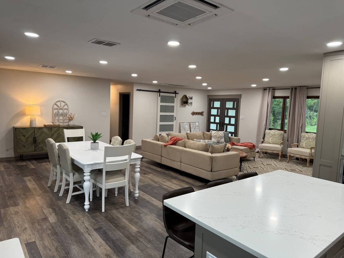 A spacious open living area is featured, with a comfortable beige sofa arranged around a central coffee table. A white dining table is positioned nearby, surrounded by chairs. Large windows allow natural light to illuminate the space, complemented by soft recessed lighting.