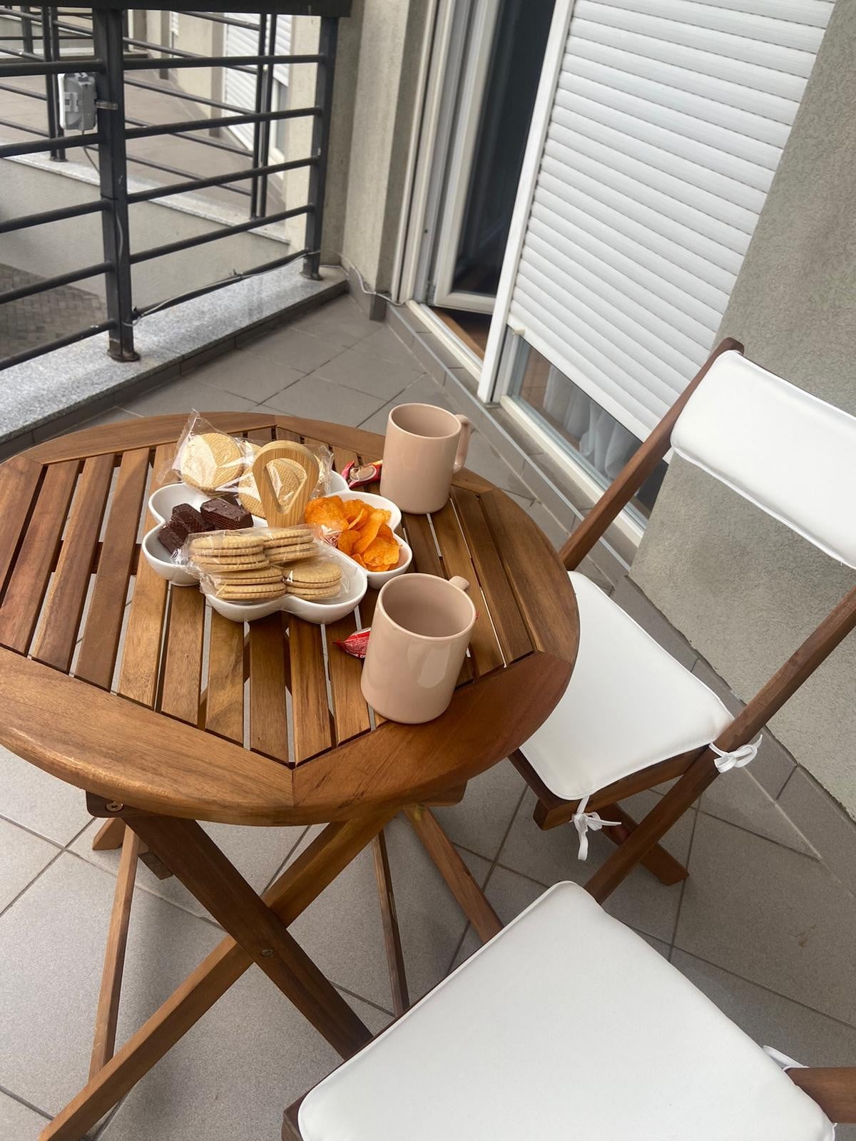A wooden table with a round top is set on a balcony, featuring a variety of pastries displayed on a decorative plate. Two beige mugs are placed nearby, alongside a folding chair with a white seat cushion. Sunlight illuminates the scene, highlighting the cozy setting.