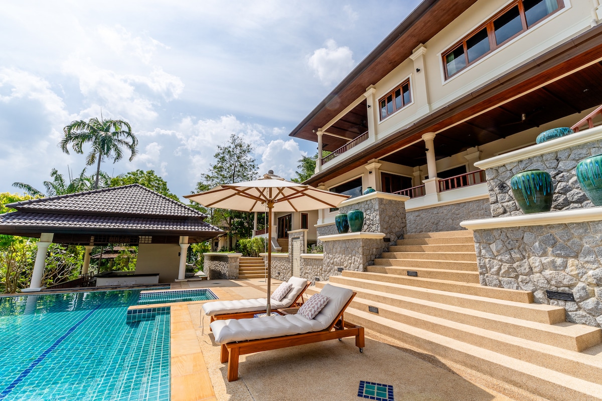 A sunlit terrace features loungers beside a shimmering infinity pool. Steps lead up to a grand villa with an expansive outdoor living area, surrounded by lush greenery. A shaded seating area is visible in the background, offering additional relaxation options.