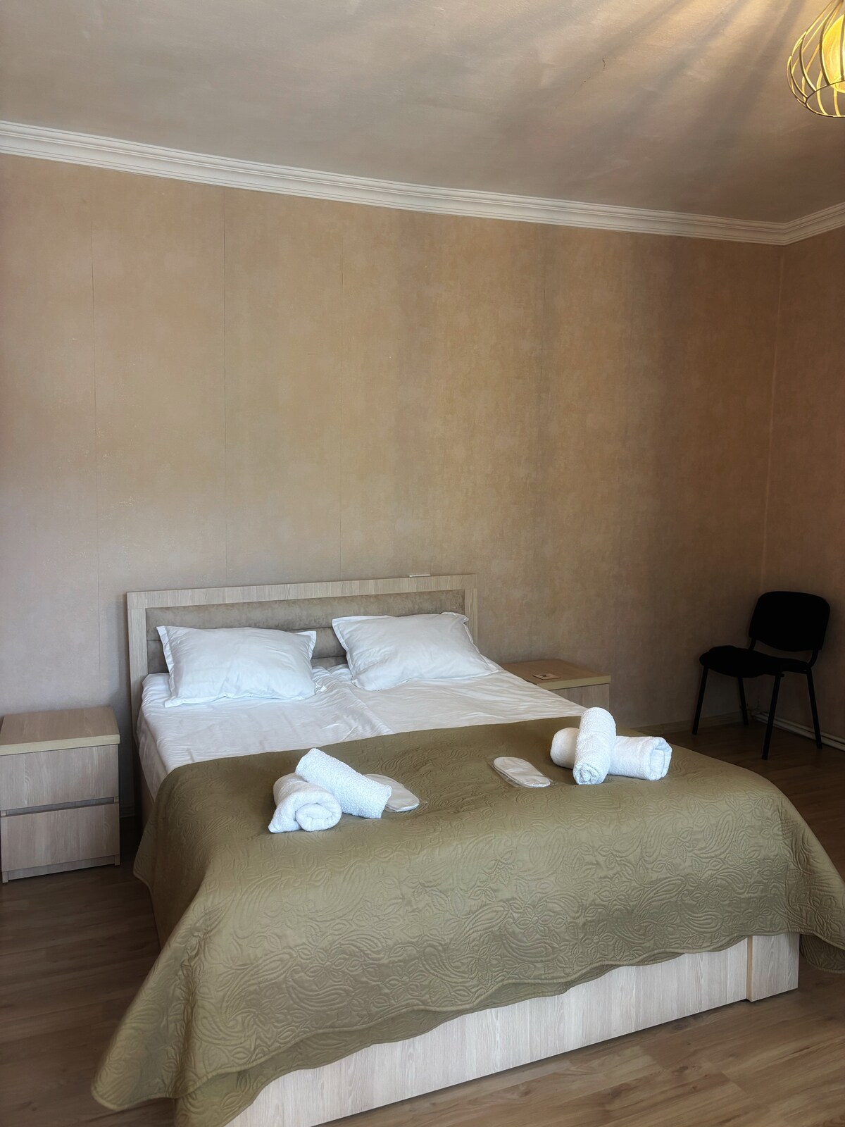 A spacious sleeping area is furnished with a bed featuring a neutral-colored quilt. Two pillows are arranged at the head of the bed. A nightstand is positioned beside the bed, and a black chair is placed against the wall, adding to the functionality of the room.