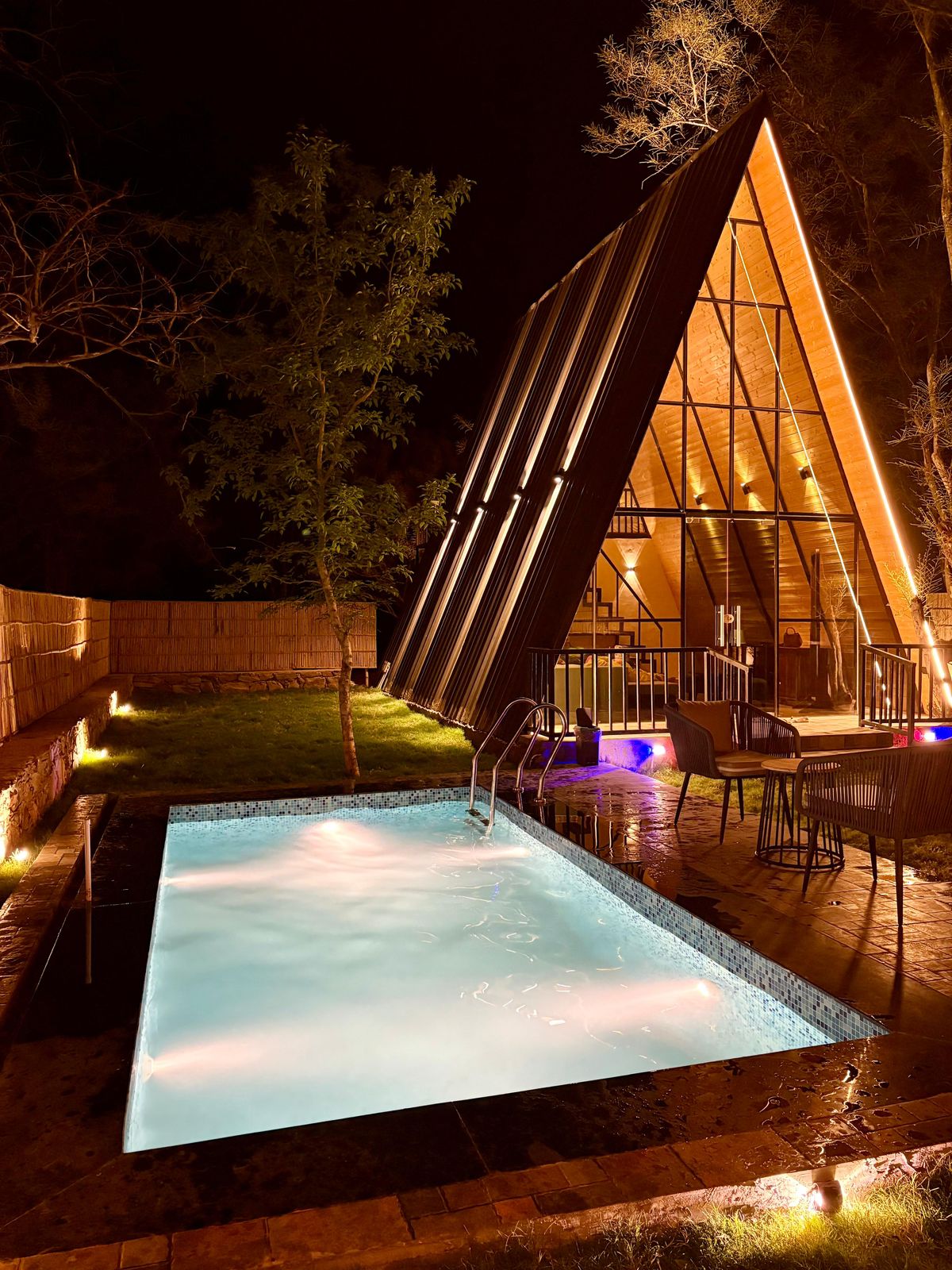 A triangular villa structure is illuminated at night, featuring large windows that showcase the interior warmth. In front, a small swimming pool reflects the surrounding lights, while a lawn area is bordered by trees, creating a serene outdoor space.