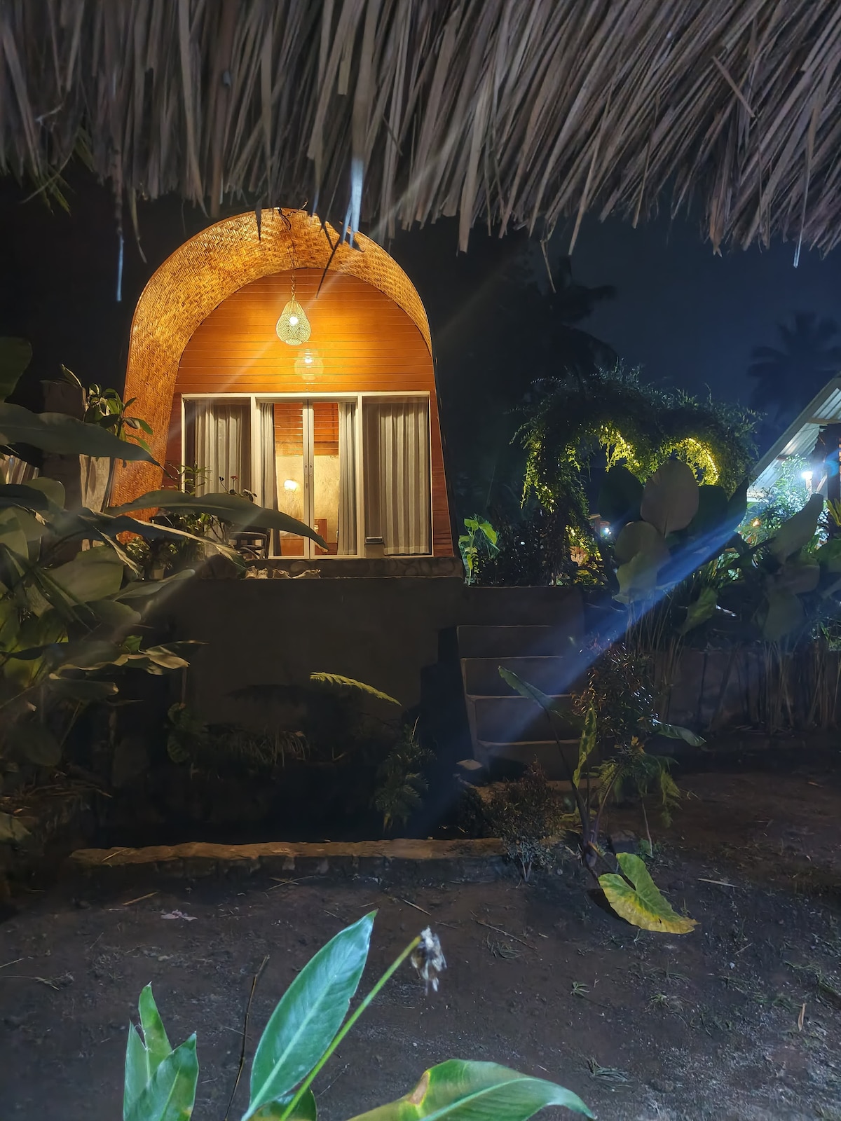 An architectural structure emerges from a lush garden, showcasing a curved entrance illuminated by a pendant light. Surrounding foliage creates a serene atmosphere, while stairs lead up to the entrance, suggesting a welcoming access point.