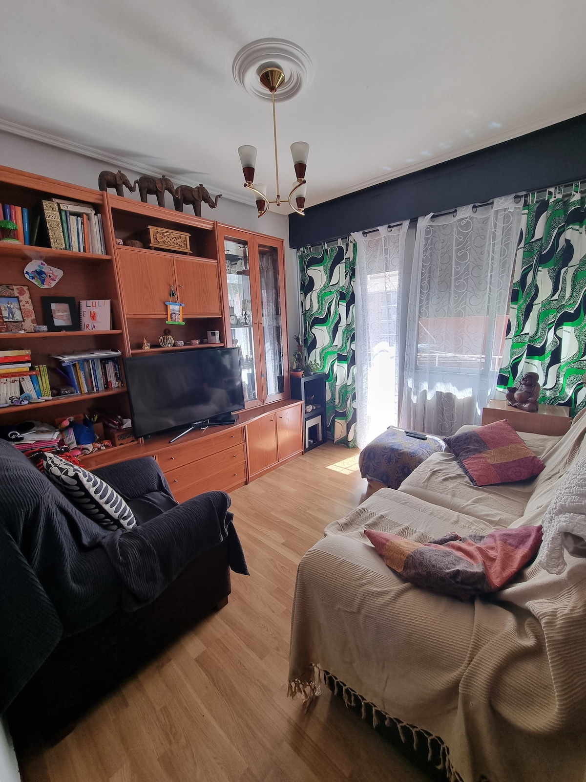 A cozy living area is featured, with a dark armchair and a light-colored sofa adorned with a woven throw. A wooden bookshelf displays various books and decorative items. Natural light enters through a large window, complemented by patterned curtains, creating a warm, inviting atmosphere.