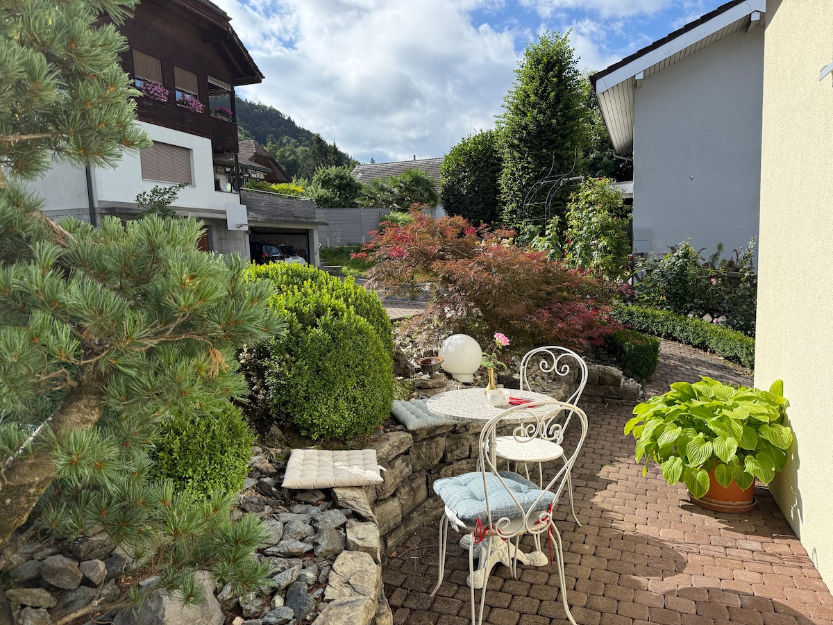 A garden area is visible, featuring neatly arranged plants, shrubs, and flowers. A small white table with a light blue cushion is positioned next to a decorative chair. Stone pathways and a variety of greenery create a tranquil outdoor space.
