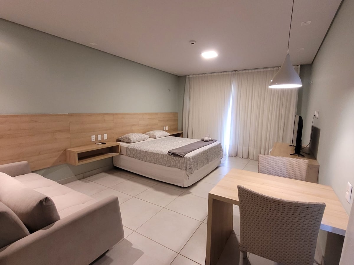 A spacious bedroom is featured with a king-size bed, a light-colored quilt, and a cozy sofa. A wooden desk and chair are placed nearby, along with a TV mounted on the wall. Soft natural light filters through sheer curtains covering a large window.