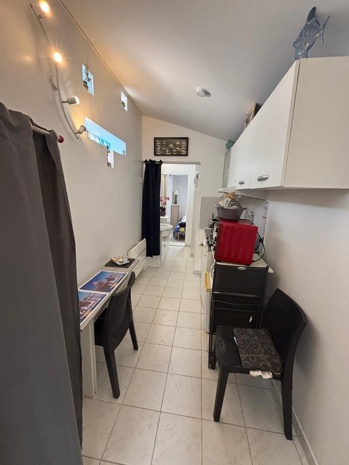 A narrow hallway is seen, featuring a compact kitchen area on one side. A small table with magazines is paired with a black chair, while a curtain provides privacy. Light from a window illuminates the tiled floor, showcasing a clean and organized space.