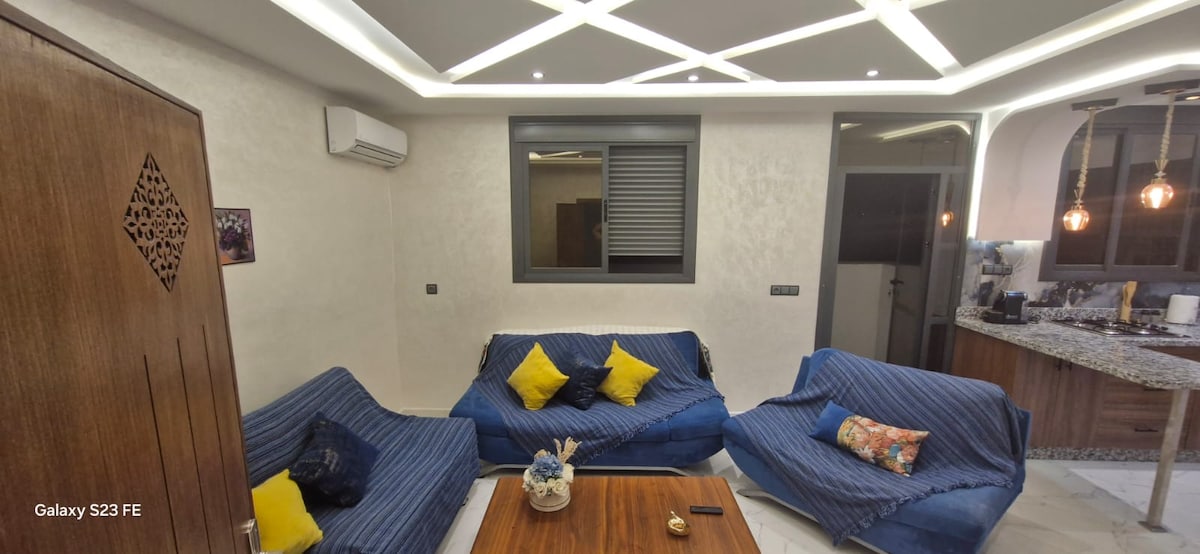 A modern living area is illuminated by warm lighting, featuring two blue sofas adorned with decorative pillows. A wooden coffee table sits centrally, complemented by a small vase. A large window allows for natural light, while the adjoining kitchenette is visible in the background.
