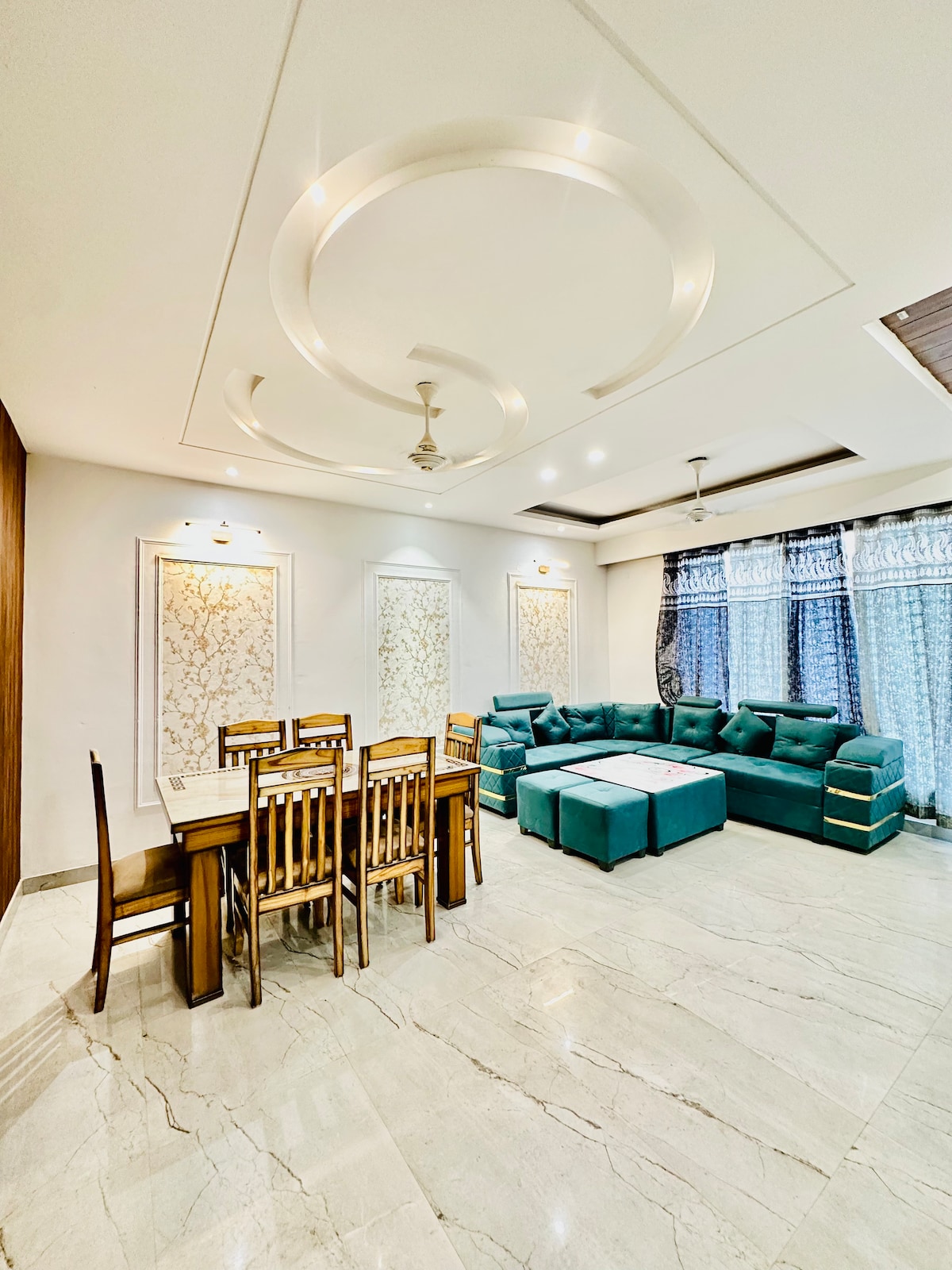 The spacious living room features a light-colored marble floor and modern ceiling design. A large, L-shaped turquoise sofa is complemented by a wooden dining table with matching chairs. Decorative wall panels add subtle elegance, while natural light filters through the sheer-curtained windows.