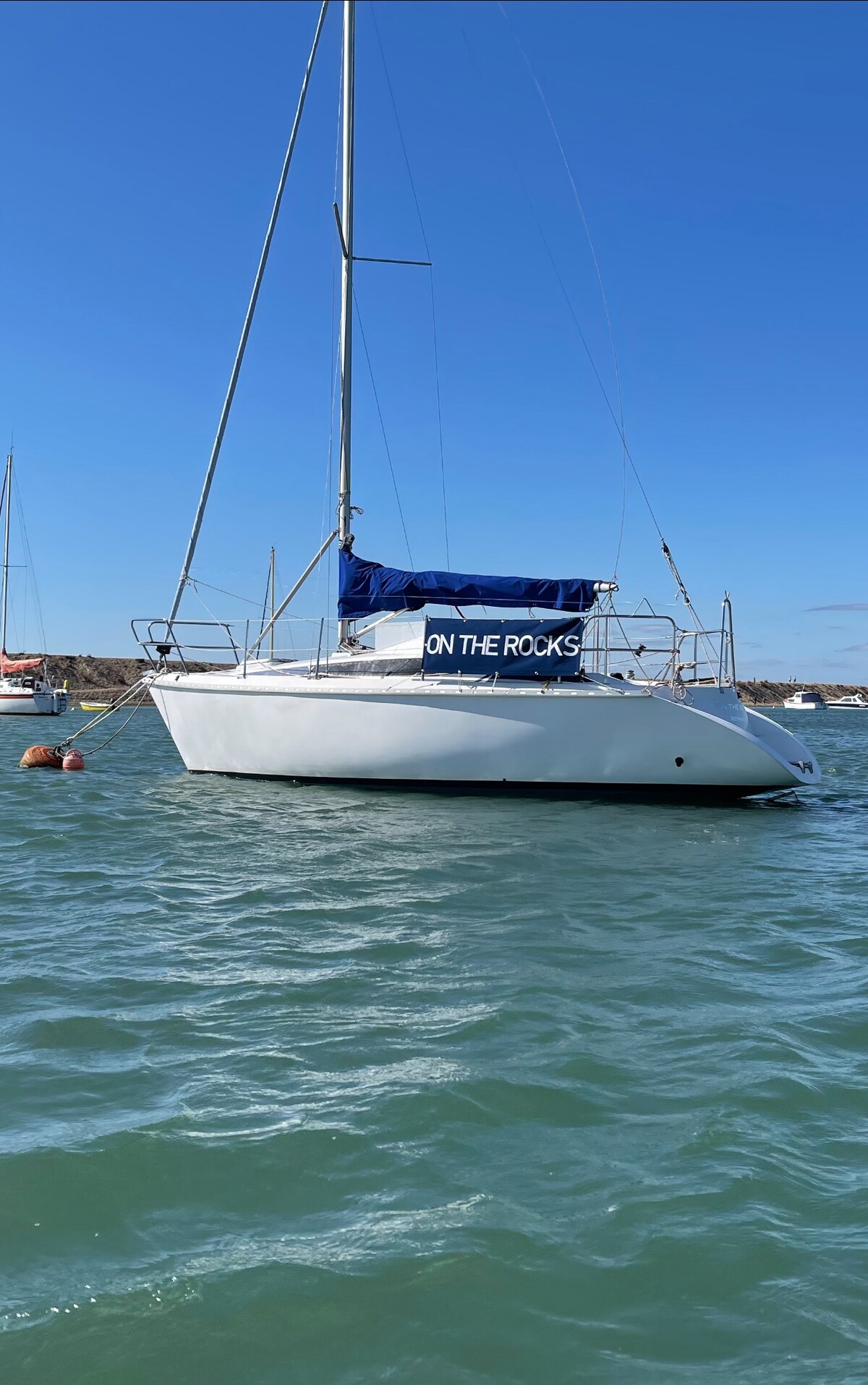 A yacht is anchored in calm waters, displaying the name 'ON THE ROCKS' on its blue canopy. The structure features a sleek, white hull and is surrounded by a clear blue sky and gentle waves. Other vessels are visible in the background.
