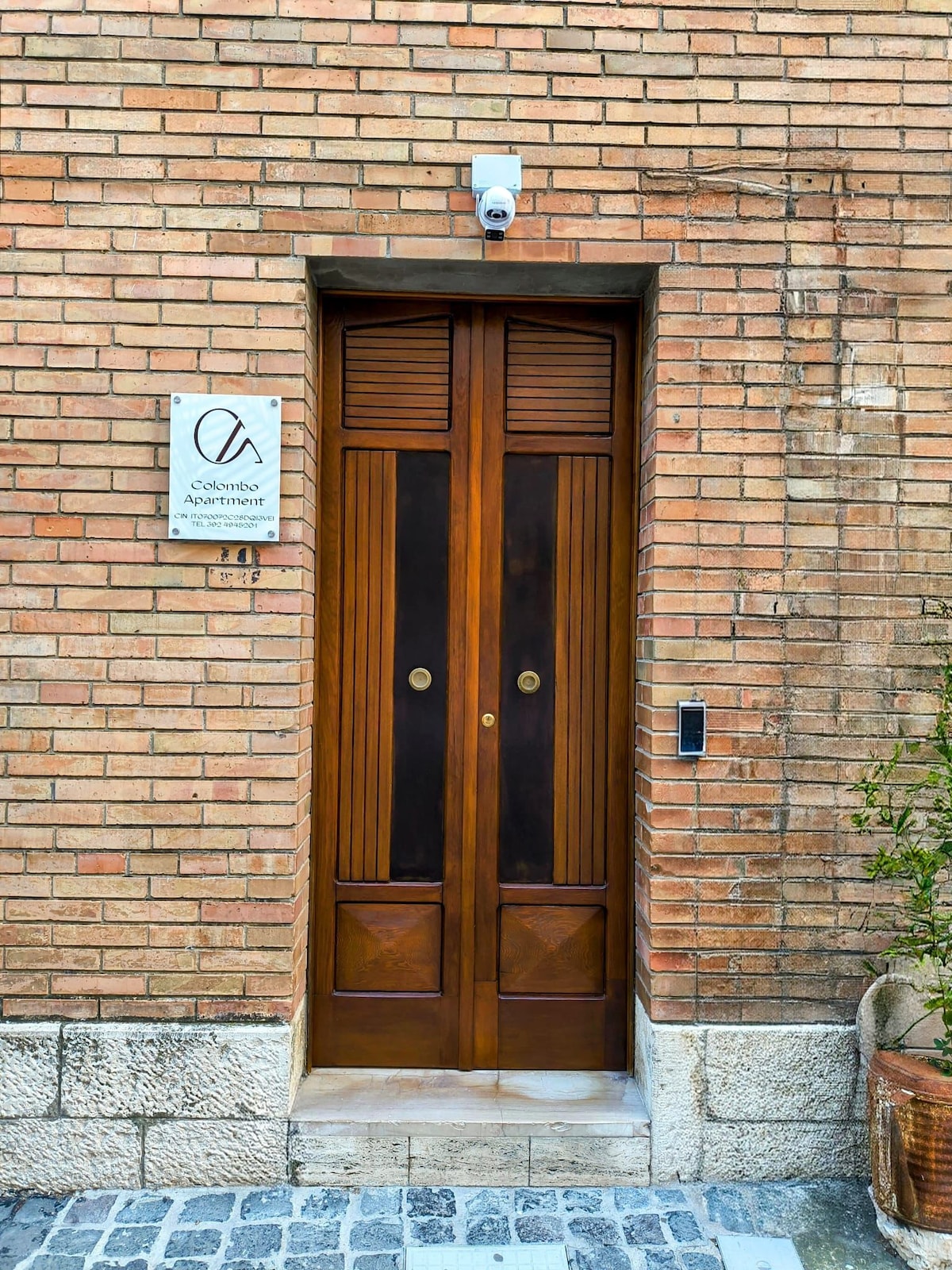 The entrance features a double wooden door with a dark finish and circular handles. A small sign with the apartment's name is displayed above the door. The surrounding brick wall provides a rustic texture, complemented by a decorative plant in a pot on the right side.