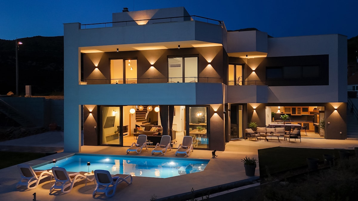 A modern two-story villa showcases a well-lit exterior at night, highlighting a swimming pool surrounded by lounge chairs. The large glass doors reveal an inviting indoor space with contemporary furnishings and warm lighting, seamlessly connecting the indoor and outdoor areas.