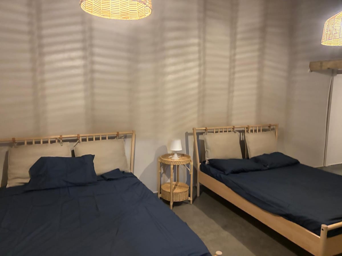 The sleeping area features two wooden beds dressed in dark bedding, providing a cozy and calm environment. A small wooden side table is positioned between the beds, topped with a simple lamp. Soft light filters through woven pendant lamps hanging above.