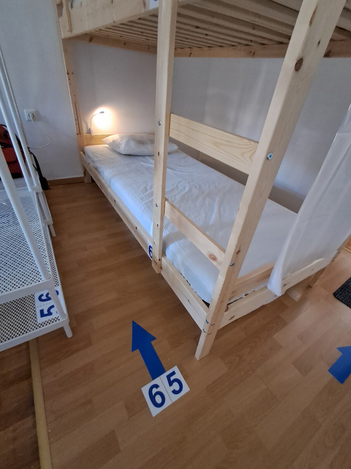 A wooden bunk bed is positioned against the wall, featuring a neatly made single bed with white linens. A bedside lamp provides soft lighting for reading. Light wood laminate flooring complements the space, which includes numbered markers on the floor for easy navigation.