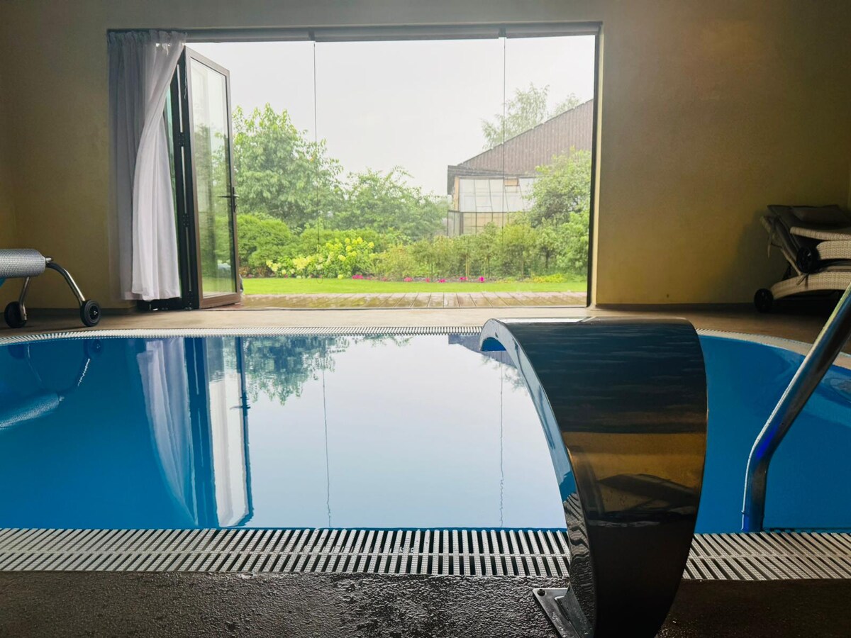 An indoor pool is featured, showcasing a sleek, modern design with a reflective surface. Large glass doors open to a lush garden area, surrounded by greenery. Daylight filters in, enhancing the serene ambiance of the space.