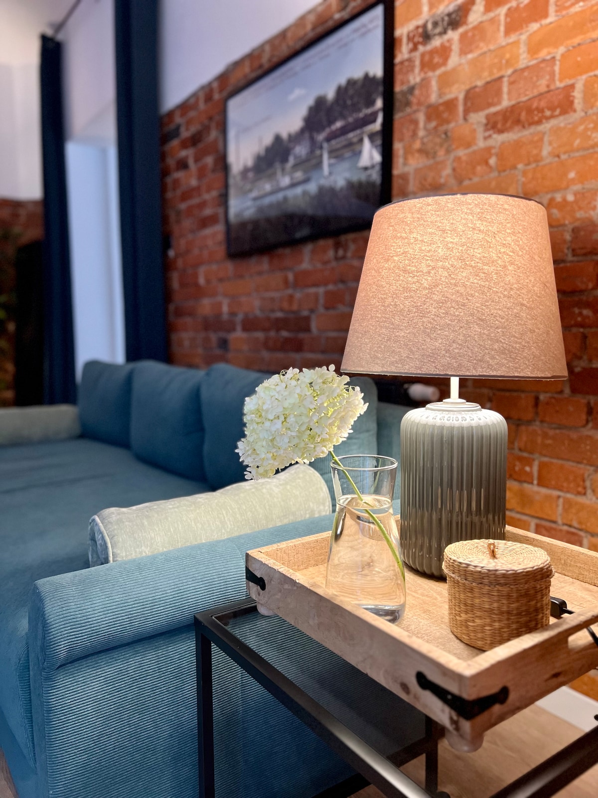 A cozy seating area features a spacious blue sofa against a backdrop of exposed brick walls. A side table holds a vase with white flowers and a decorative lamp, providing soft lighting. A framed artwork depicting a serene landscape is displayed above.