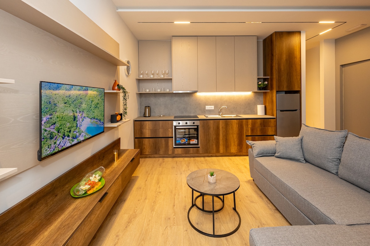 A modern kitchen and living area are showcased, featuring a sleek cabinetry design in warm wood tones. A comfortable gray sofa is positioned around a circular coffee table. A flat-screen television is mounted on the wall, while overhead lighting enhances the space's functionality.