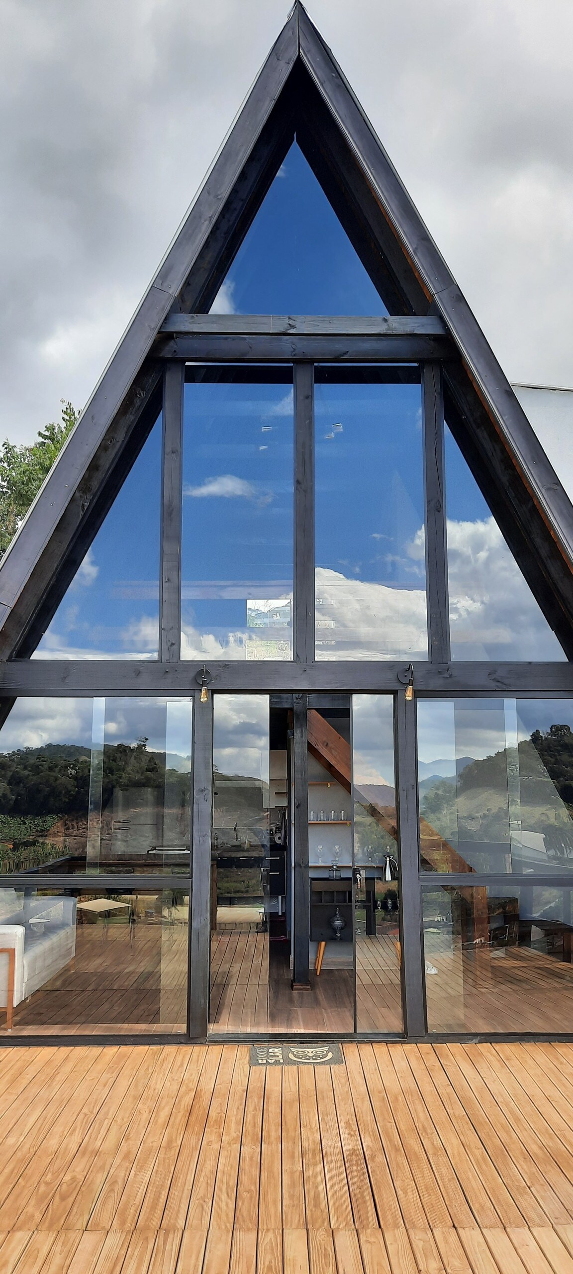 The exterior of the chalé features a striking A-frame design with large glass panels that reflect the surrounding landscape. A wooden deck extends in front, offering a seamless transition between indoor and outdoor living spaces. The sky is visible through the glass, enhancing the airy feeling.