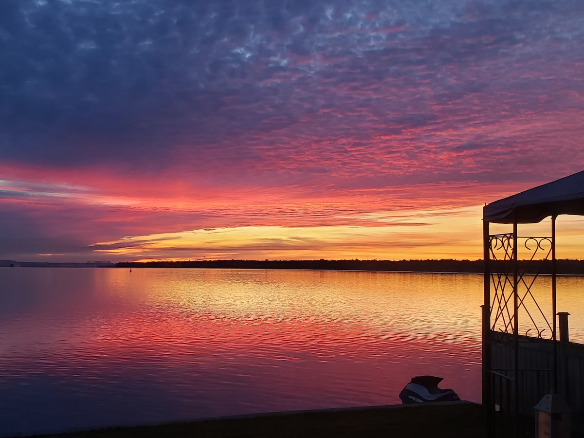 A vibrant sunset casts shades of pink, orange, and purple over calm waters. The sky reflects on the surface, creating a serene scene. A structure with a decorative frame is visible in the foreground, complementing the natural beauty of the landscape.