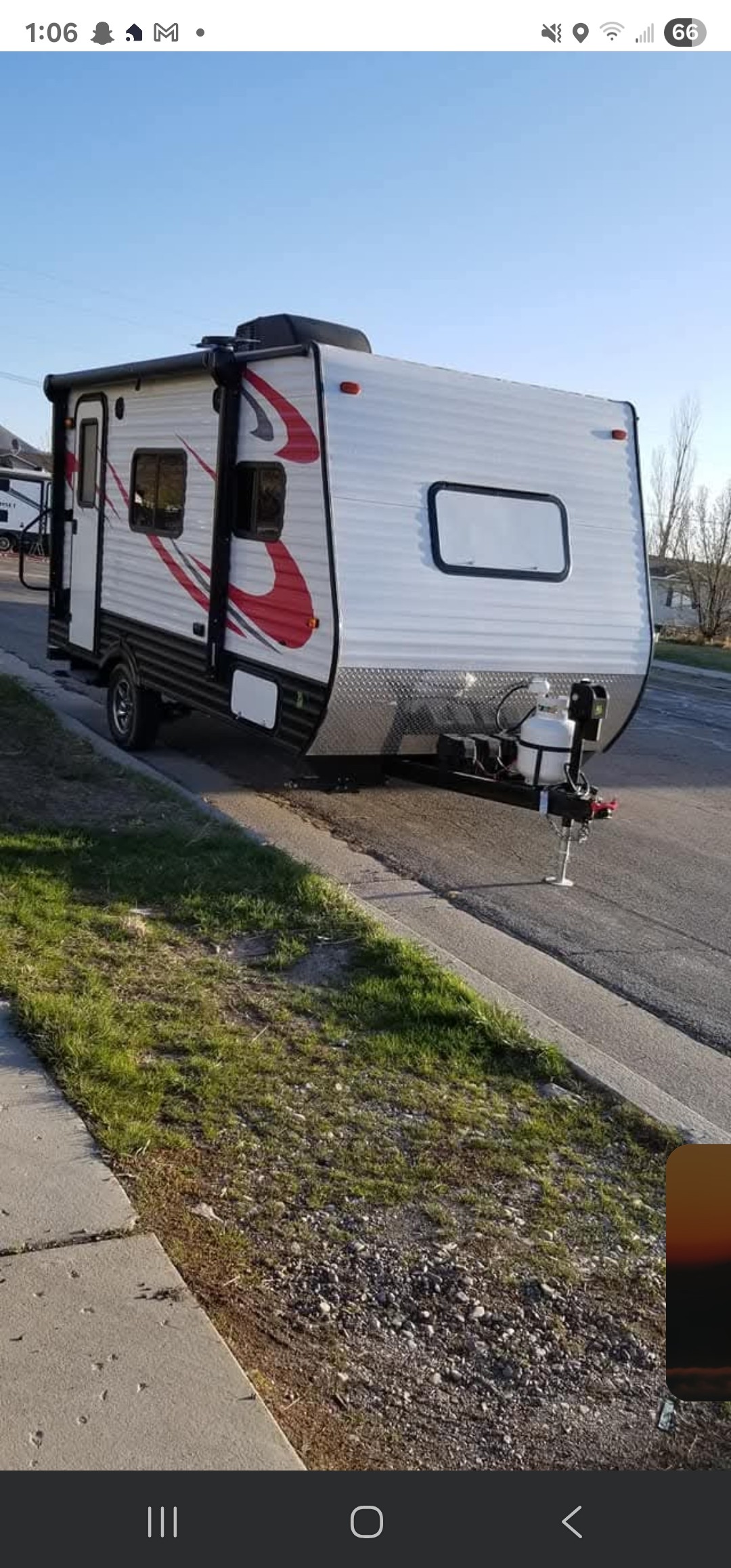 A compact trailer sits on the side of a suburban street, showcasing a sleek exterior with a combination of silver and red detailing. The environment features a grassy area in the foreground, with paved road visible beside the trailer.