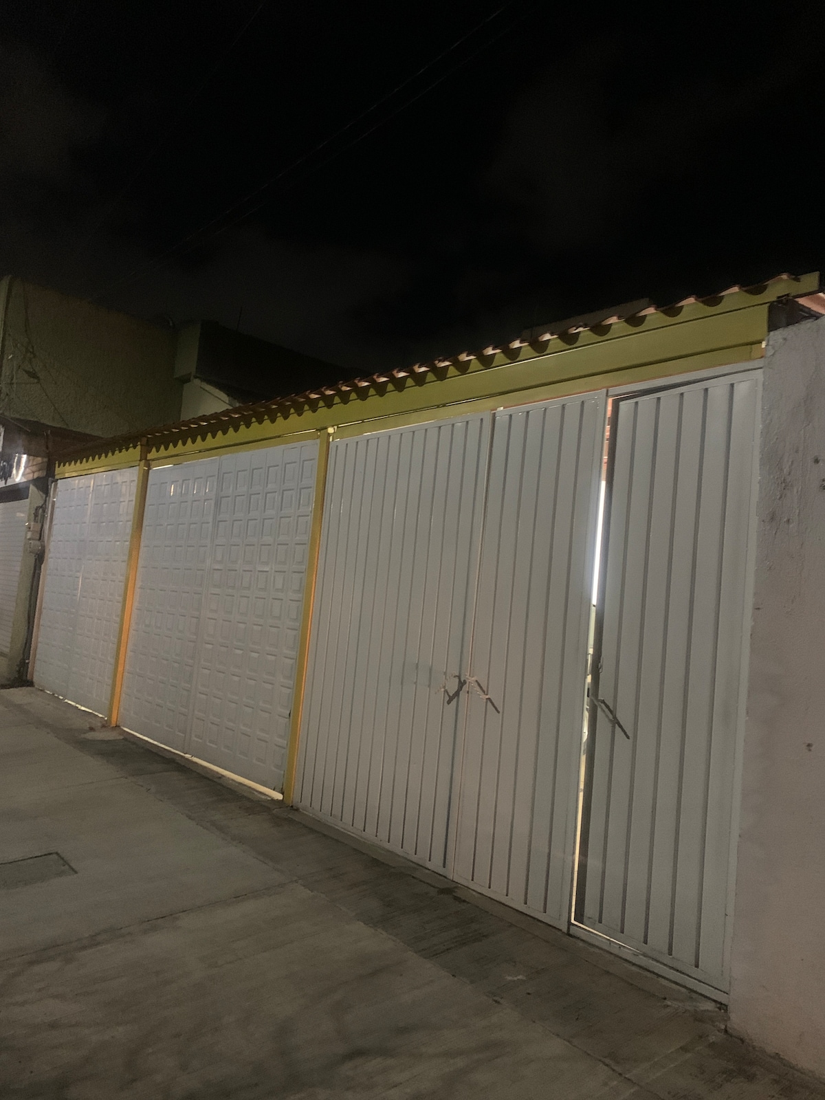 A large sliding gate with white and yellow elements creates a secure entrance. The gate is partially open, revealing a glimpse of the space beyond. The surrounding area is dimly lit, highlighting the structure against the night sky.