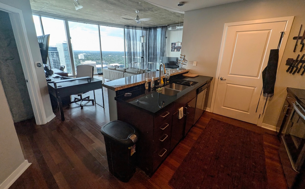 A modern kitchen area features sleek dark cabinetry and a granite countertop with a double sink. A small wastebasket is positioned beside the cabinetry. Natural light fills the space through floor-to-ceiling windows, while a workspace with a desk is visible in the background.