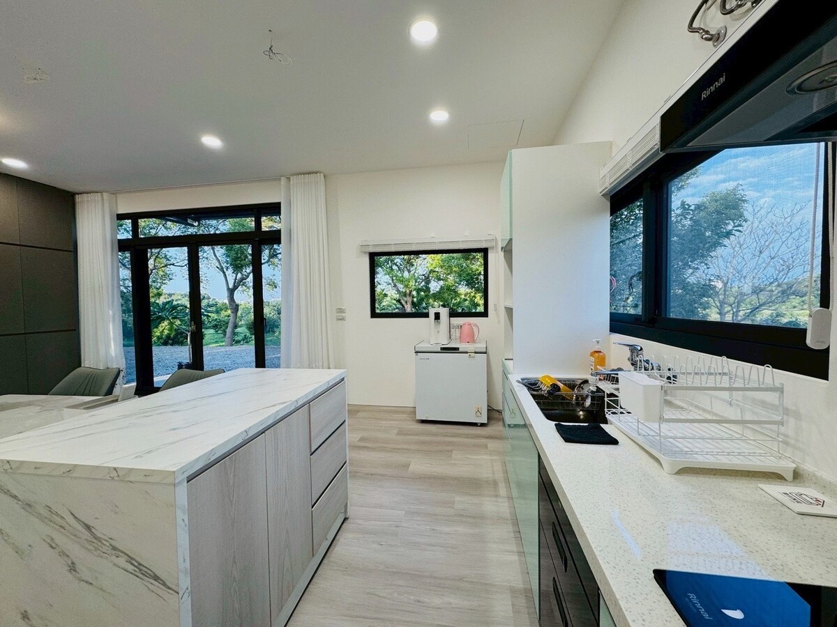 A modern kitchen area features a white countertop and a marble island, complemented by dark cabinets. Large windows invite natural light and offer views of greenery outside. Essential appliances, including a refrigerator and sink, are easily accessible. Stylish seating is arranged nearby.