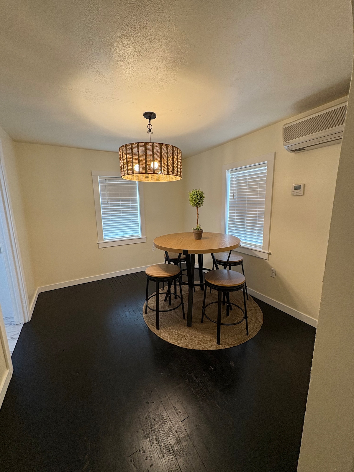 A small dining area features a round wooden table surrounded by three tall chairs. A potted plant is centered on the table under a contemporary light fixture. Two windows allow natural light, complemented by a smooth, dark wooden floor.