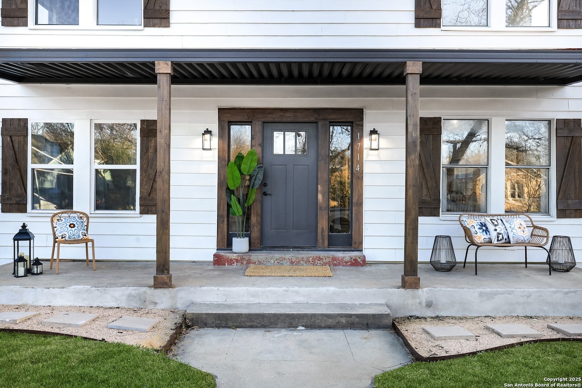 An exterior view showcases a welcoming entrance with a modern front door framed by two large windows. Two chairs are positioned on either side of the doorway, accompanied by potted plants. A decorative mat is placed at the entrance, enhancing the inviting appearance.