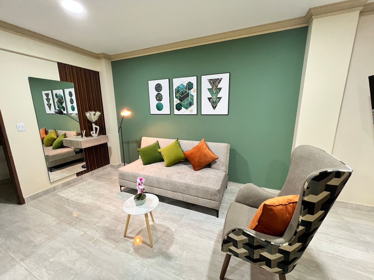 A modern living area is presented, featuring a light brown sofa adorned with green and orange cushions. A small round table, topped with an orchid, sits in front of the sofa. Two geometric art pieces are displayed on a green accent wall, enhancing the space's visual appeal.