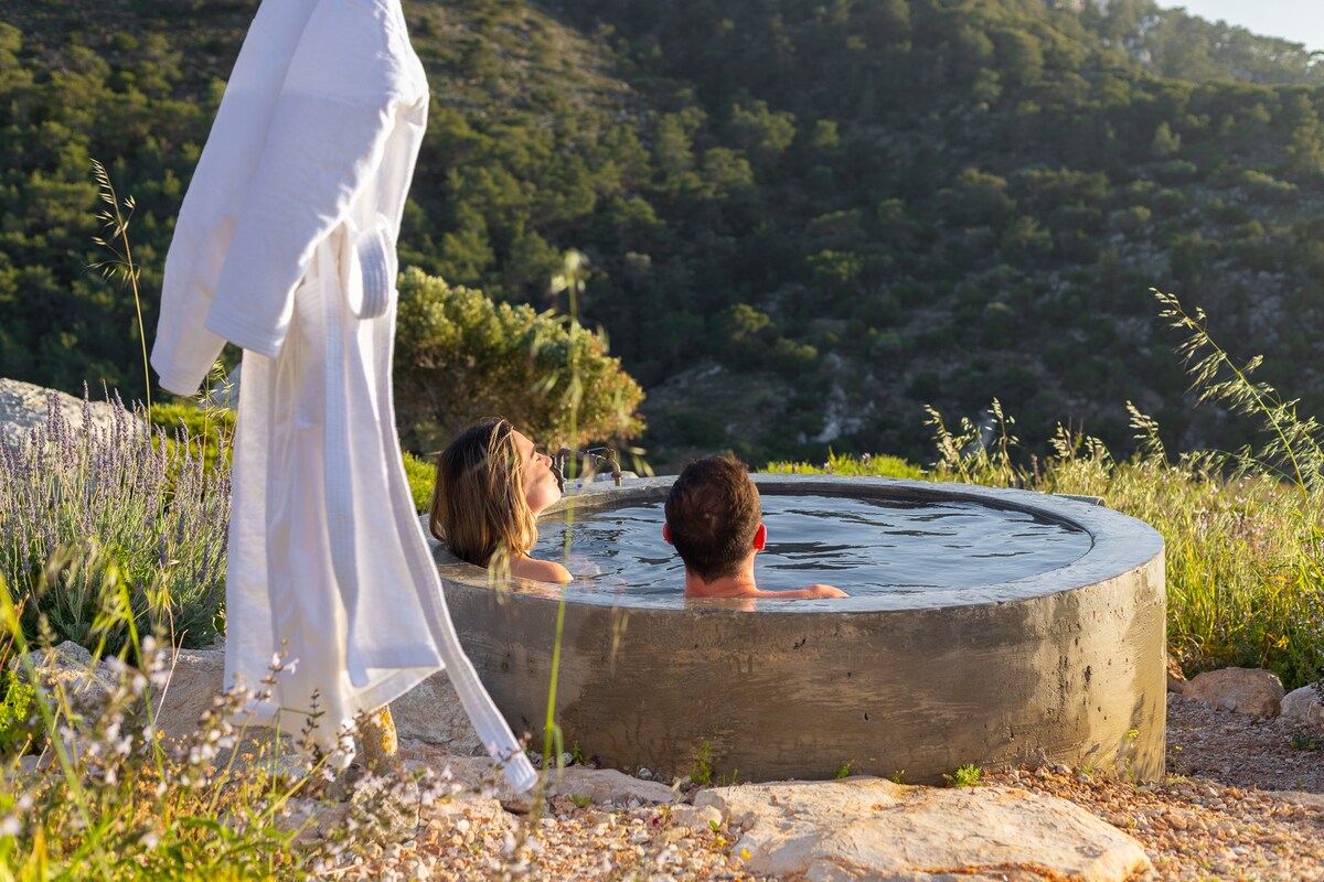 An outdoor tub, surrounded by natural vegetation, is featured in a serene setting. Two guests are seen relaxing in the water, with a lush, green hillside visible in the background. A white robe hangs nearby, suggesting comfort and ease in this tranquil escape.