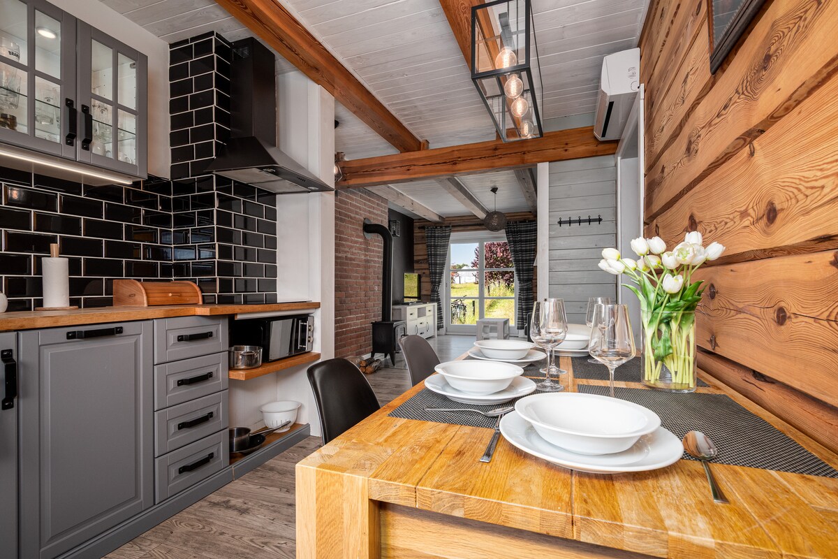 A well-equipped kitchen area is complemented by wooden beams and a mix of light and dark cabinetry. A dining table is set for four, featuring white plates and glassware, with a vase of fresh tulips adding a touch of nature. A window offers views into the surroundings.