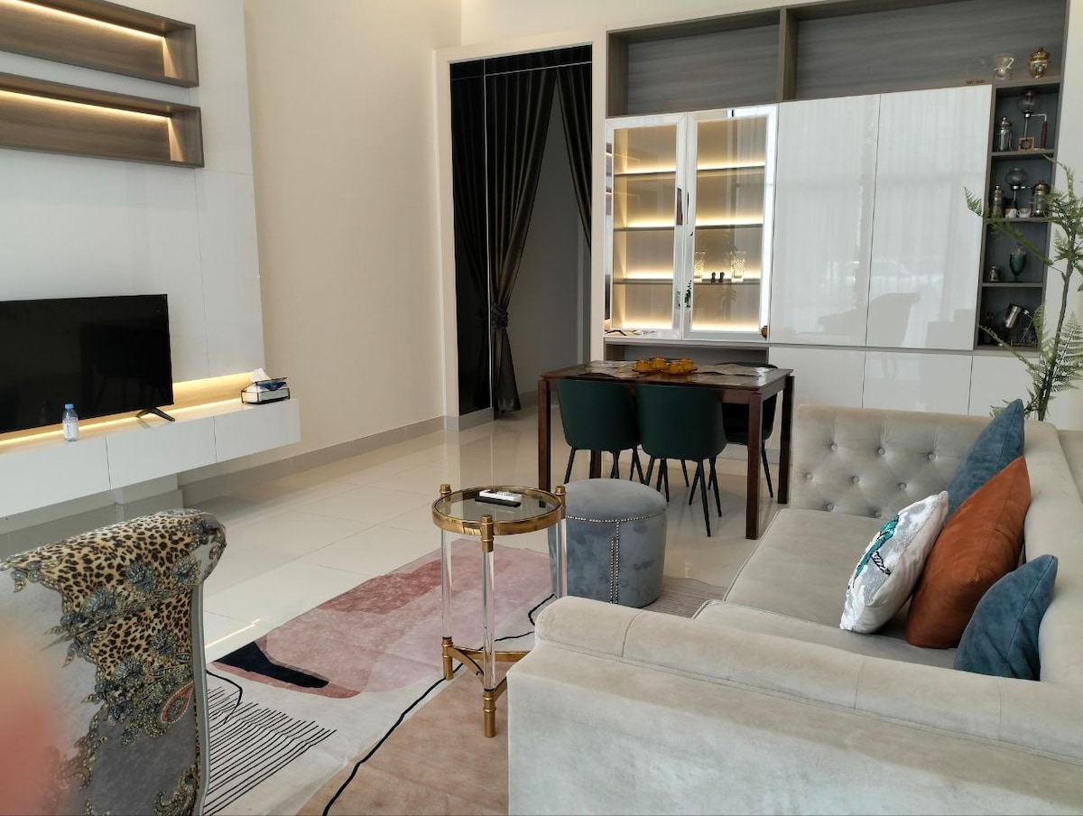 A modern living area is showcased, featuring a comfortable beige sofa and a decorative accent chair with a unique pattern. A glass coffee table rests on a soft area rug, while a dining table with green chairs is visible in the background. Natural light fills the space.